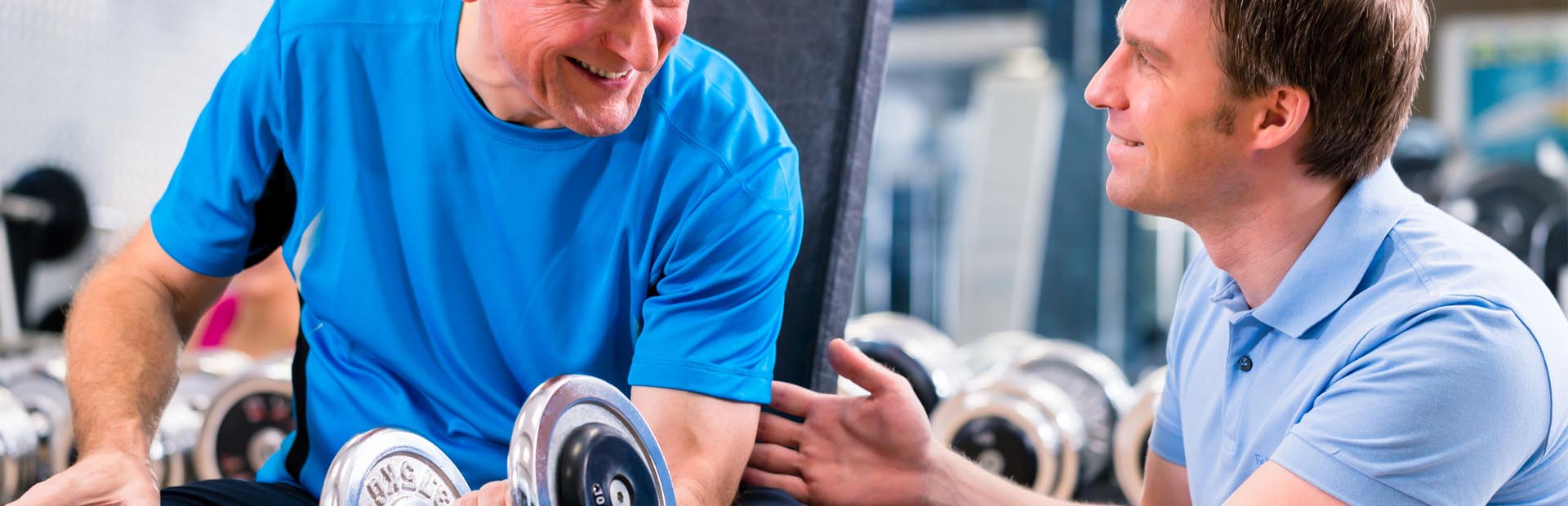 Older adult in gym holding dumbbell smiling at fitness instructor