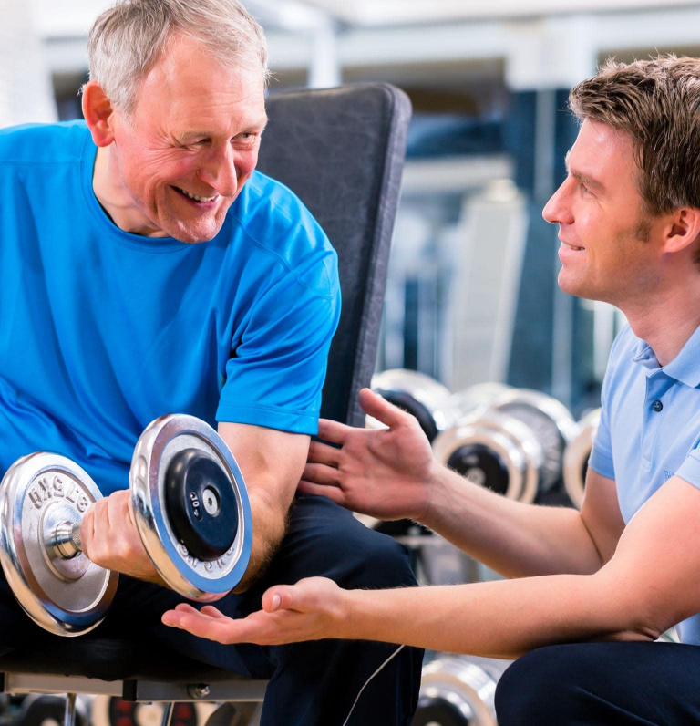 Older adult in gym holding dumbbell smiling at fitness instructor