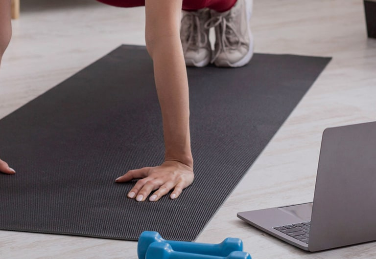 A person on a training mat with a laptop and dumbells at their side.