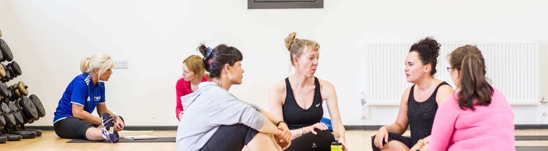 A HFE Pilates course teacher speaks to a student (left) and a circle of four students sit in discussion (right).