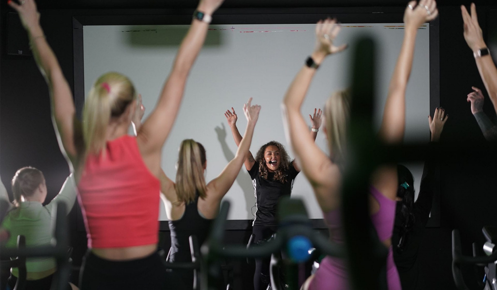 A cycling instructor at the front of a class on a cycle machine with her hands in the air while smiling and speaking into a headset. Students can be seen facing the instructor and copying her movements.