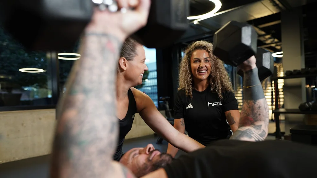 A PT guiding a student with a client completing a dumbbell bench press