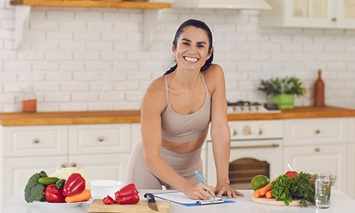 A lady in a kitchen writing a nutrition meal plan on paper and a clipboard.