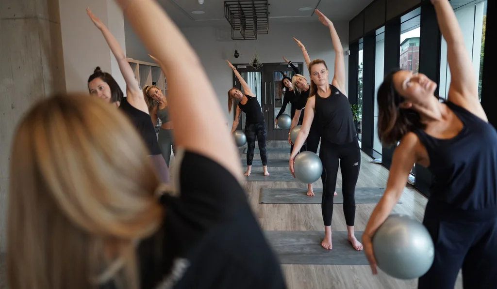 Students in a Pilates class holding a Pilates ball in one hand by their leg, with the other hand extended over their head.