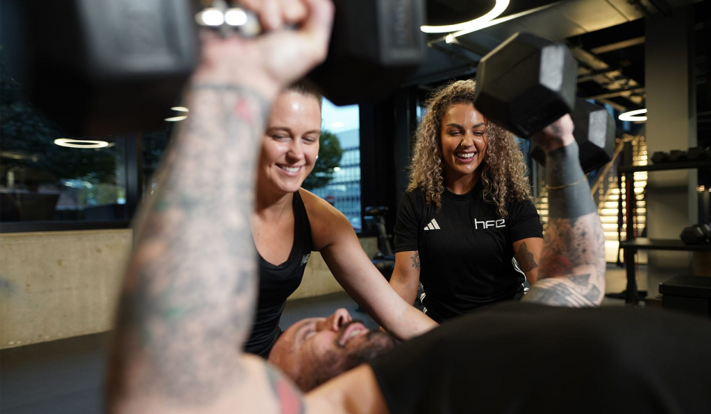 A personal trainer coaching a student, who is supporting a male to complete a bench press