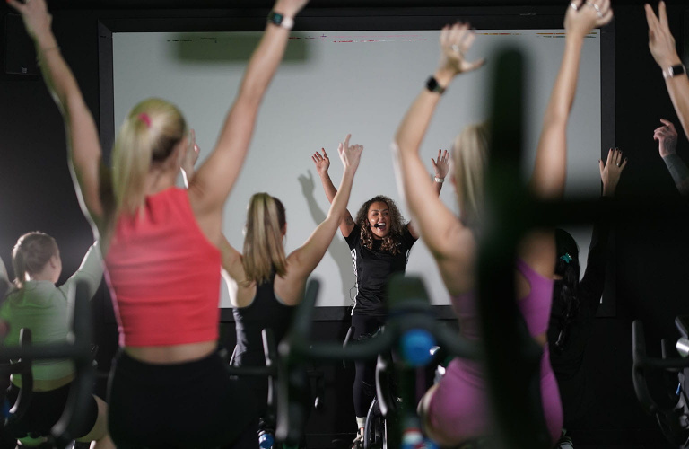 A cycling instructor at the front of a class on a cycle machine with her hands in the air while smiling and speaking into a headset. Students can be seen facing the instructor and copying her movements.