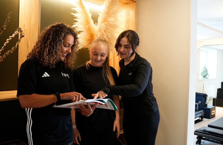 Three females in a gym studio looking at a manual together