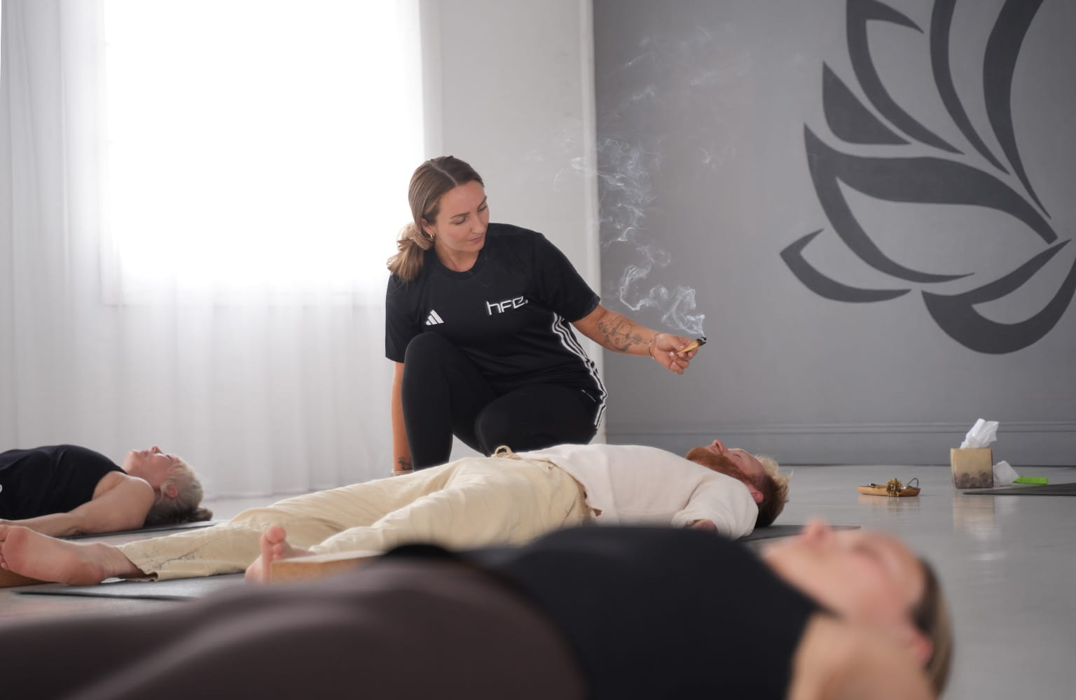 Students lying on their mats in a yoga class while a yoga teacher burns incense. 
