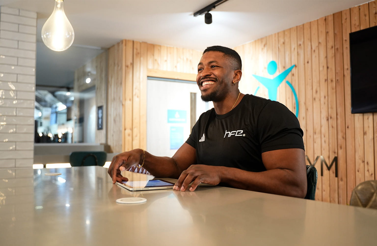 A fitness coach sat on a high table in a gym smiling