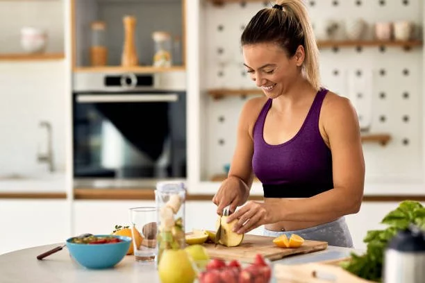 photograph of a woman smiling and chopping an apple. there is other fruit scattered across the worktop and a smoothie maker beside her.