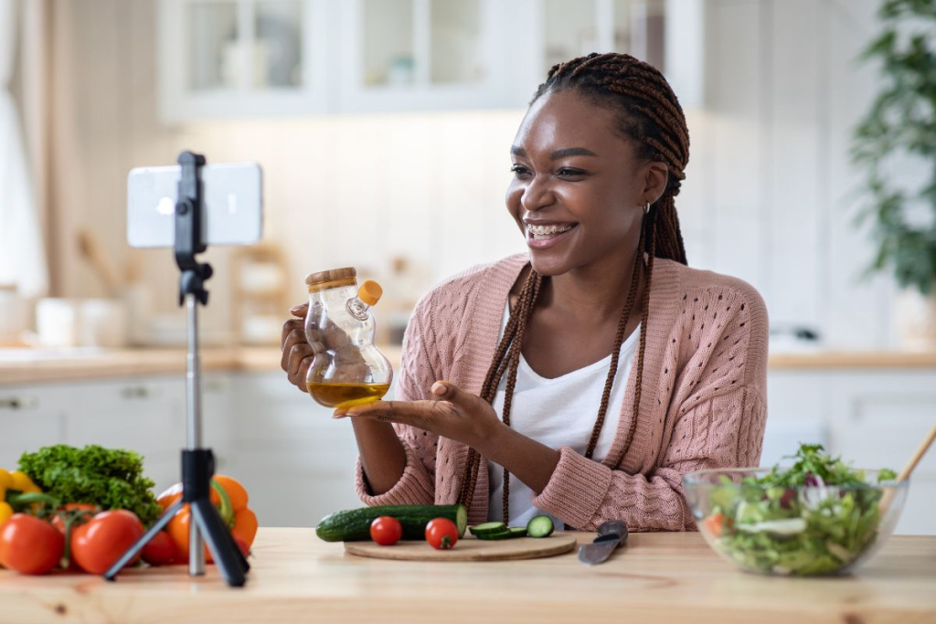 This image shows a woman sat at a kitchen table in front of her phone, which is set up on a miniature tripod. She is holding up a glass pouring bottle filled with olive oil and is smiling at her phone camera. Sher has a chopping board with a knife, chopped cucumber and tomatoes in front of her, a glass bowl full of salad on her left and a heap of broccoli, tomatoes and peppers opposite her.